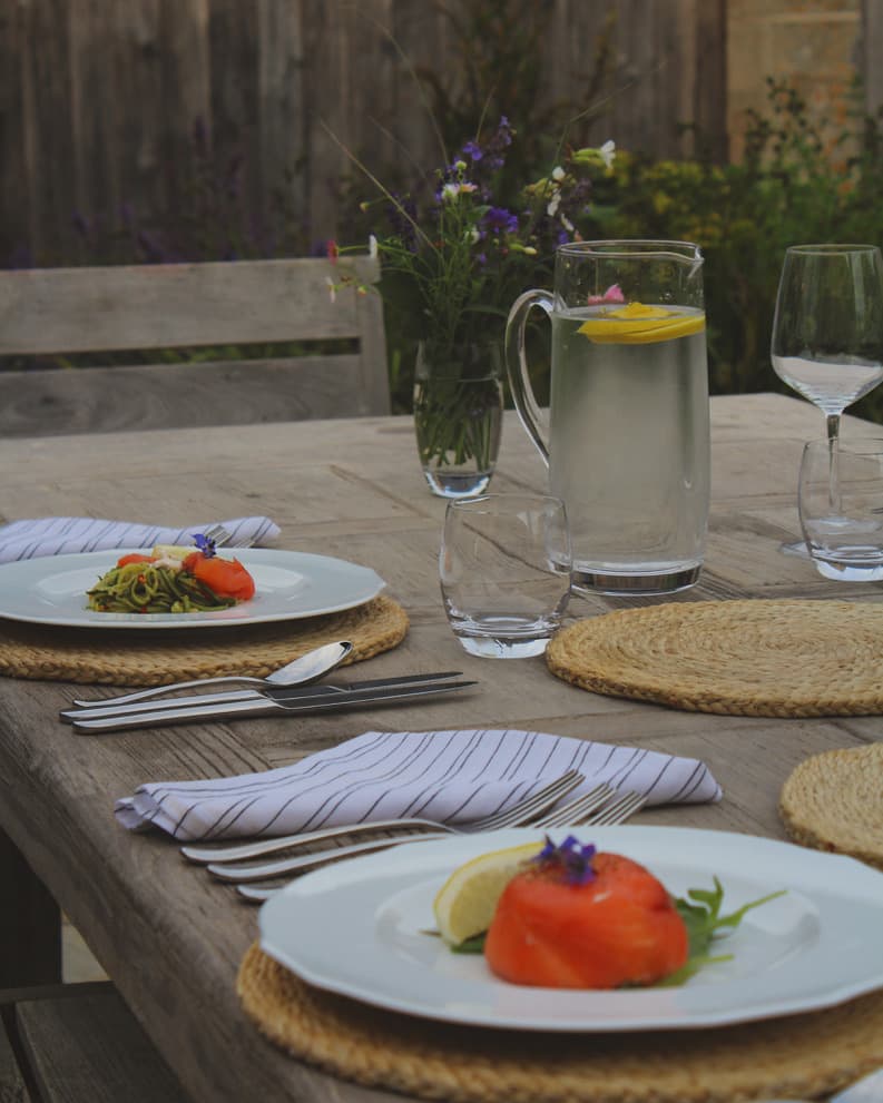 An outdoor dining setup featuring a fresh salmon appetizer on white plates, surrounded by rustic woven placemats and floral table decor.