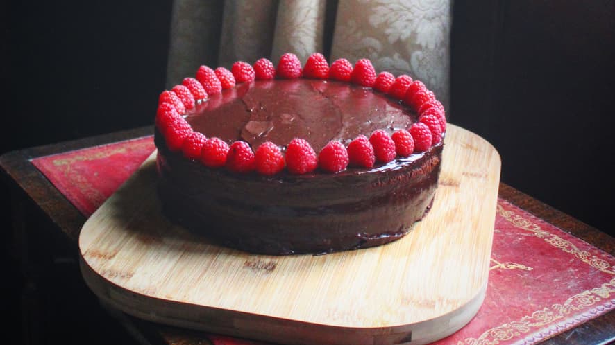 A rich chocolate cake with glossy ganache, topped with a ring of fresh raspberries, placed on a wooden board near a vintage window with soft curtains.
