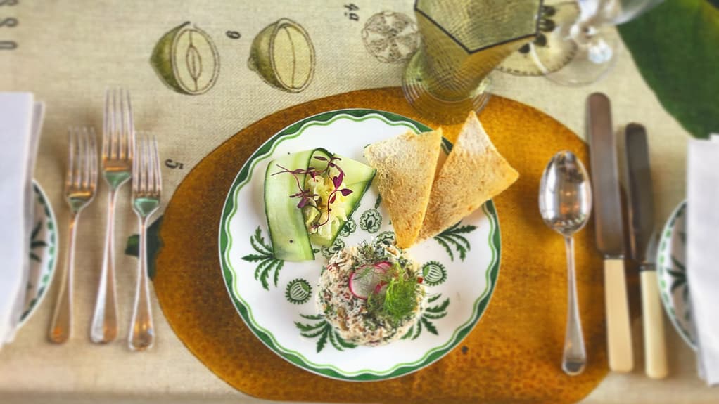 A refined plate of crab salad garnished with microgreens, served with cucumber ribbons and crisp toast on a beautifully set table with vintage silverware.