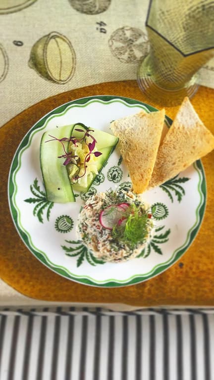 A refined plate of crab salad garnished with microgreens, served with cucumber ribbons and crisp toast on a beautifully set table with vintage silverware.