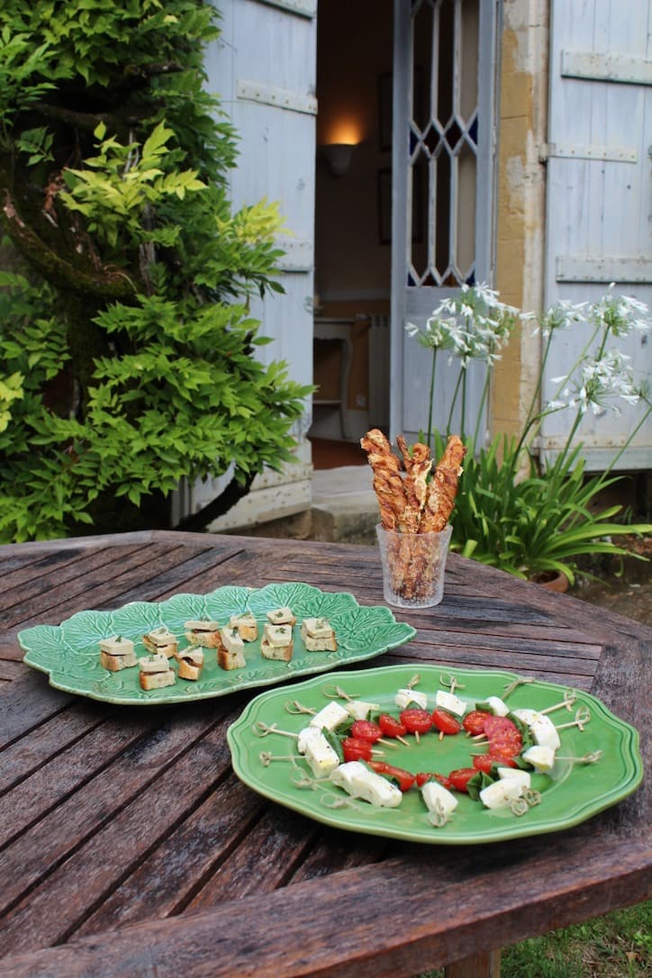 A wooden table with green plates holding caprese skewers, herbed cheese cubes, and crispy breadsticks outside a charming country house.