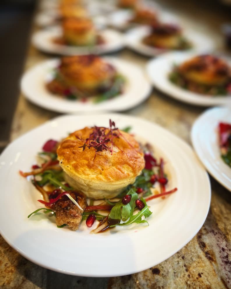 A golden brown mini pie plated on a bed of fresh arugula, drizzled with balsamic glaze and garnished with pomegranate seeds and microgreens, served at a formal dining event.