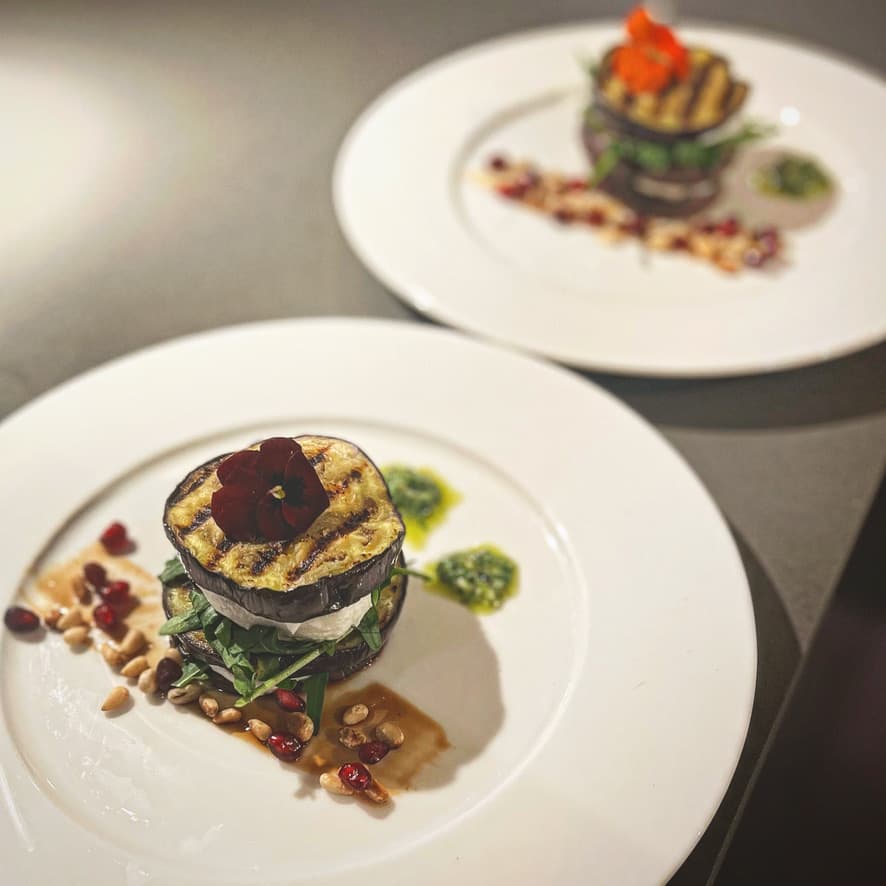 A gourmet dish featuring grilled eggplant layered with greens and cheese, elegantly plated with pesto drizzle and pine nuts.