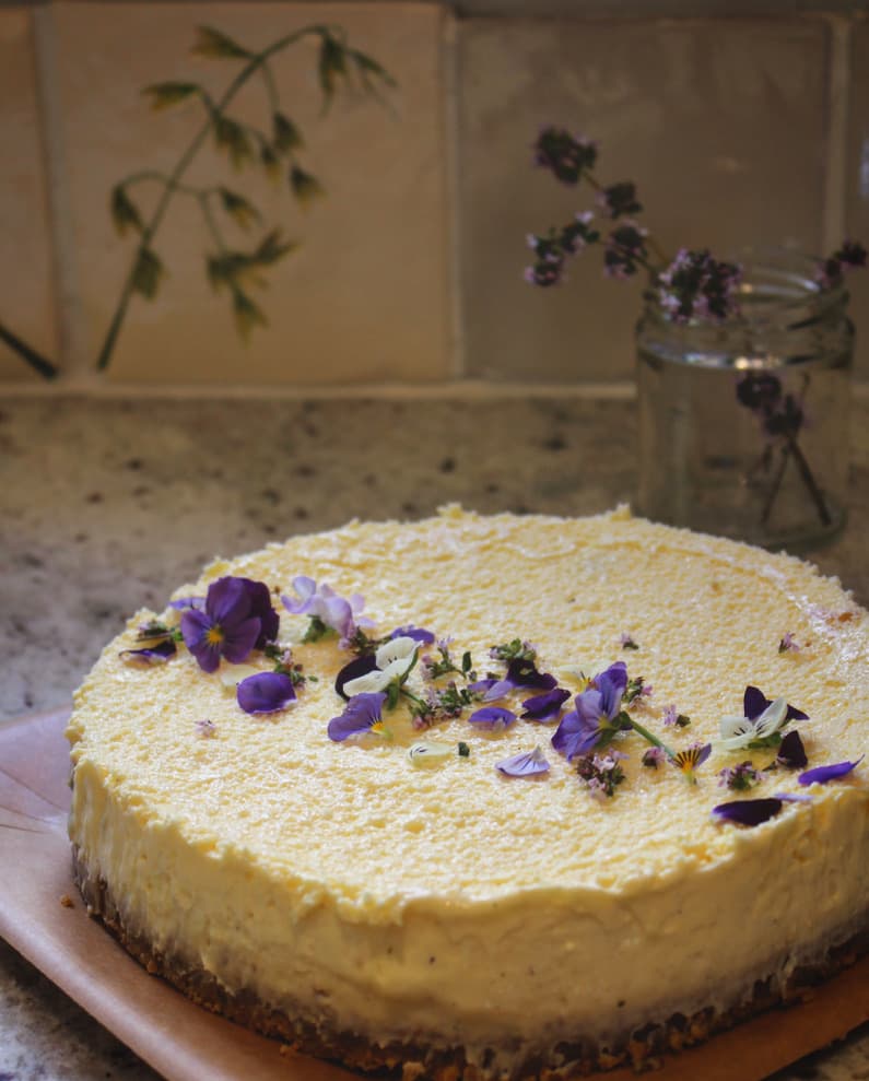 A creamy lemon cheesecake adorned with delicate edible flowers, sitting on a rustic countertop with herbs in the background.