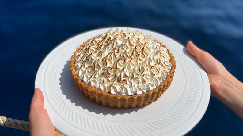 A lemon meringue pie with golden peaks, held on a white plate against a stunning blue sea and rocky coastline backdrop.