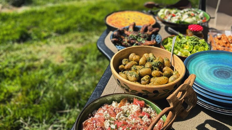 A farm-style outdoor buffet with fresh salads, potatoes, and grilled meats served in wooden and ceramic bowls on a sunny countryside backdrop.