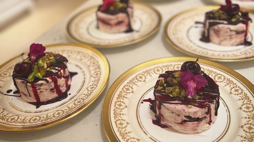 Delicate cherry mousse cakes drizzled with berry sauce, topped with pistachios and cherries, served on gold-rimmed plates in an elegant setting.