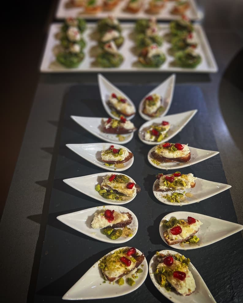 Small white spoon-shaped plates holding creamy cheese bites topped with pomegranate seeds, pistachios, and honey drizzle.