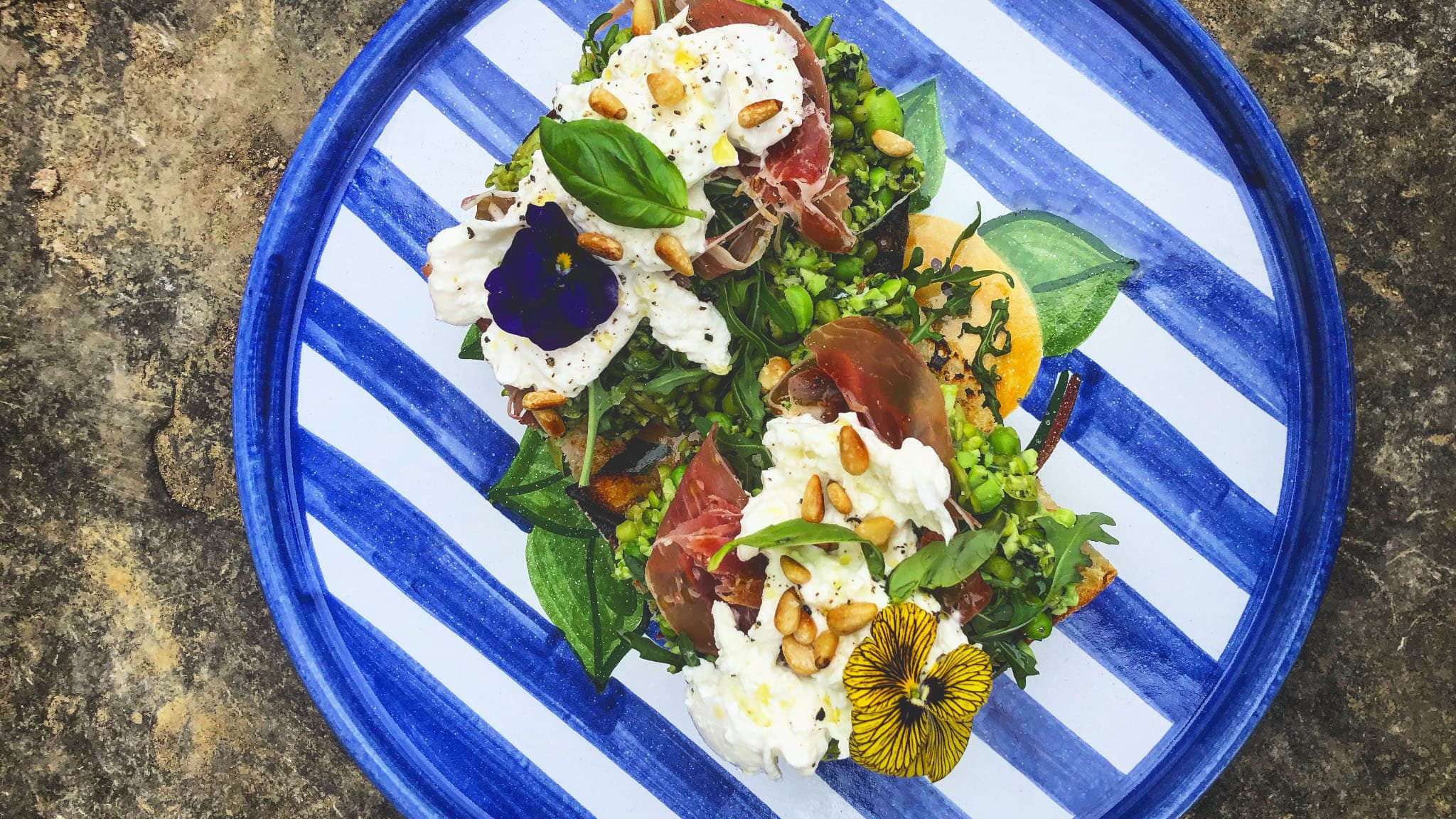 A vibrant plate of toasted bread topped with prosciutto, creamy burrata, arugula, pine nuts, and edible flowers, served on a blue striped ceramic dish.