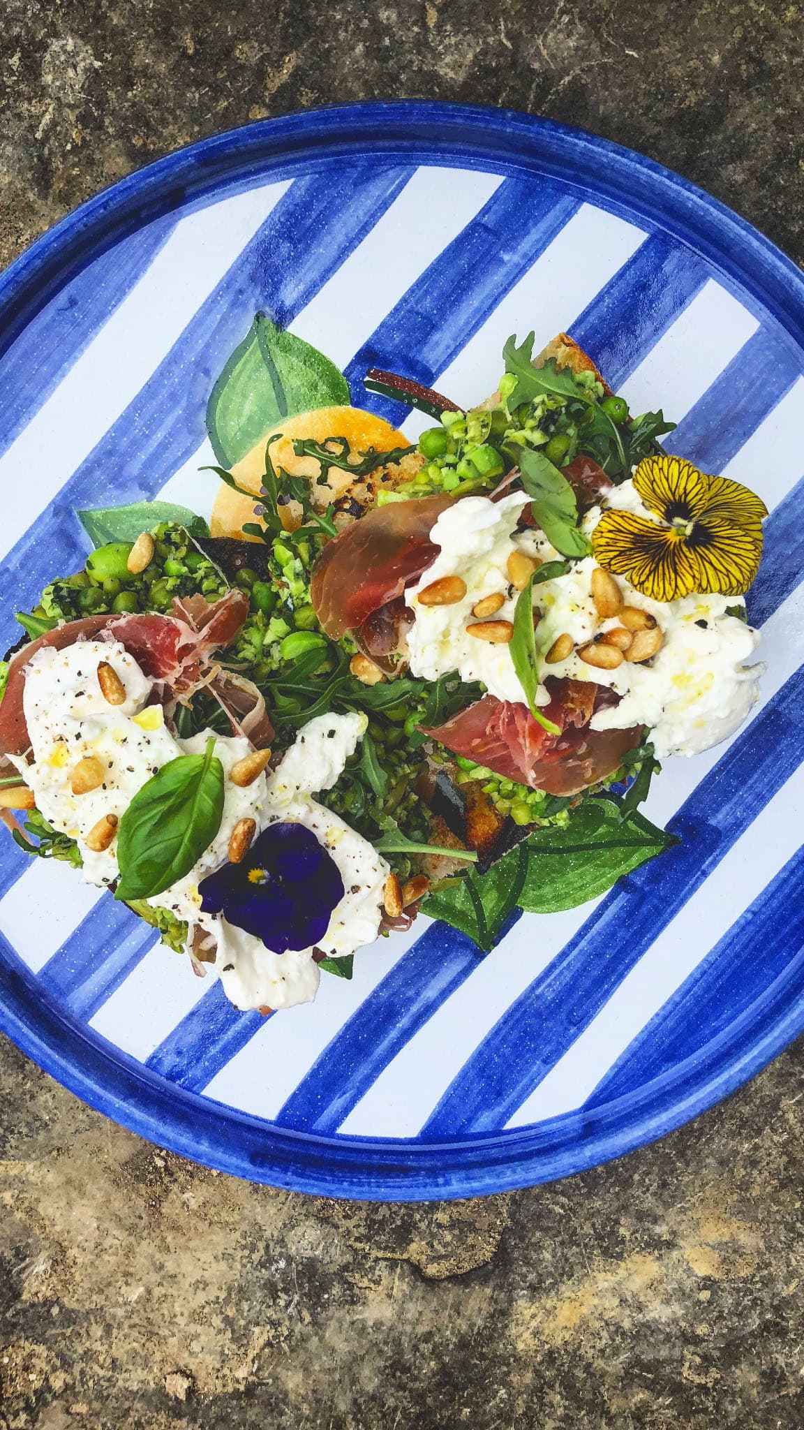 A vibrant plate of toasted bread topped with prosciutto, creamy burrata, arugula, pine nuts, and edible flowers, served on a blue striped ceramic dish.