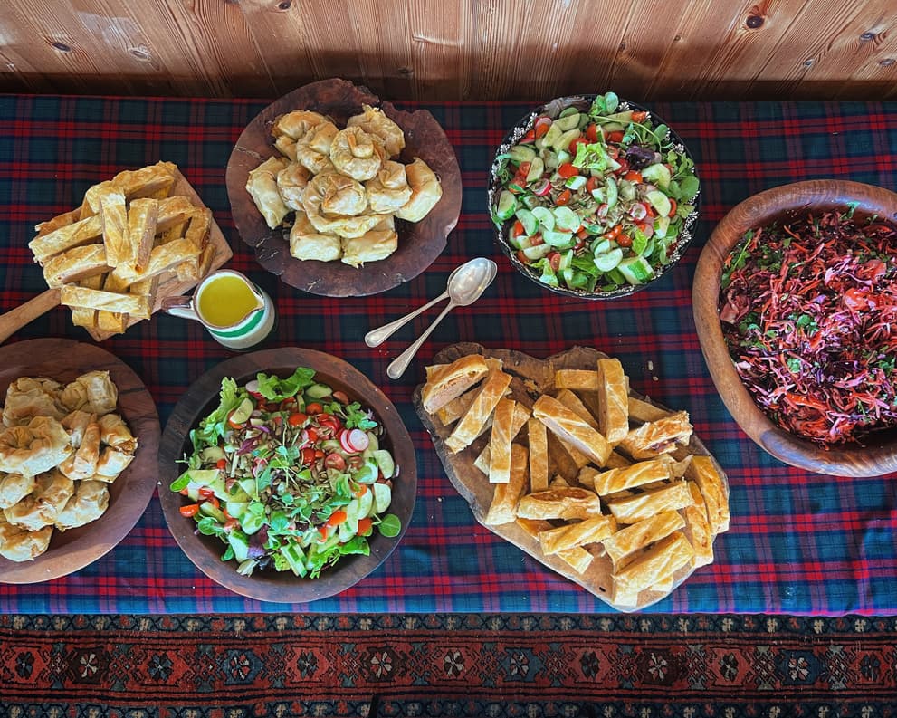 A rustic buffet spread on a tartan tablecloth with wooden serving bowls filled with fresh salads, pastries, and traditional baked goods.