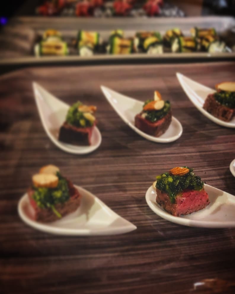 Elegant seared beef bites topped with a green herb salsa and almonds, served on modern white spoons for a stylish appetizer presentation.