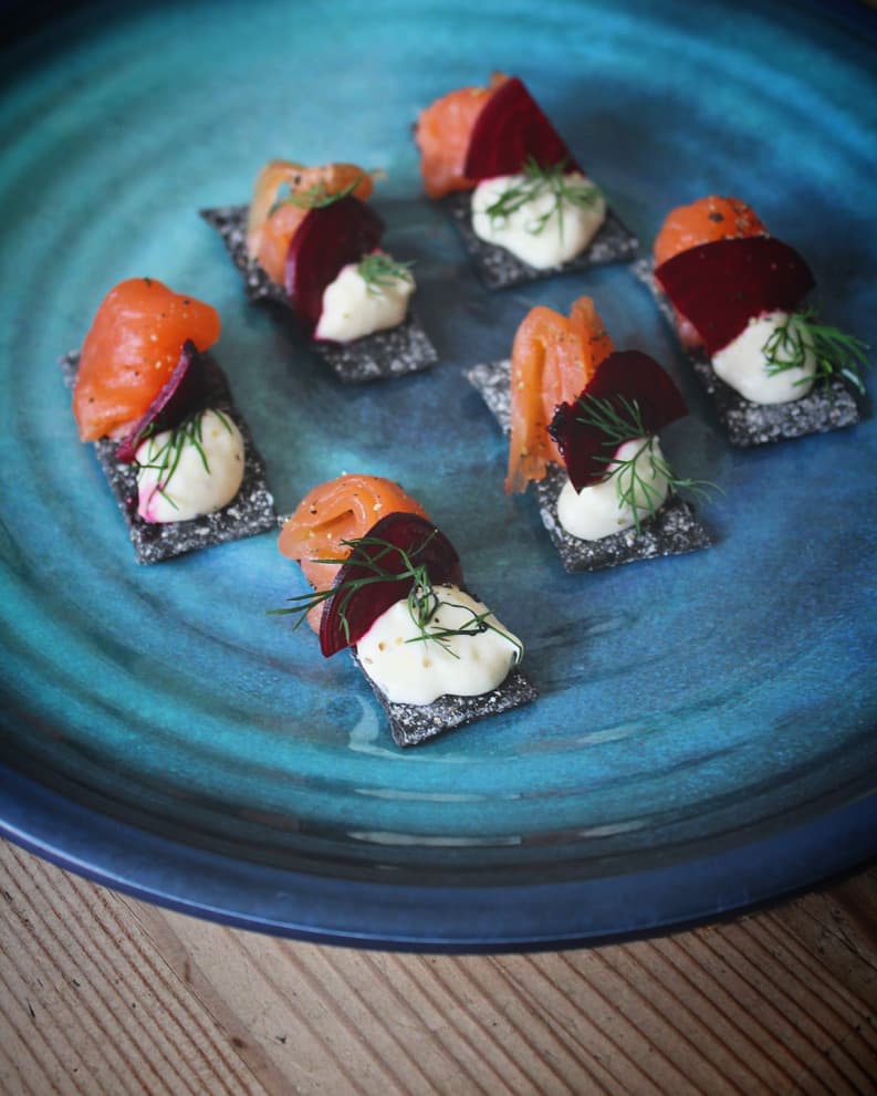 Elegant appetizers with smoked salmon, beetroot, and cream on black sesame crackers, garnished with fresh dill, arranged on a deep blue ceramic plate.