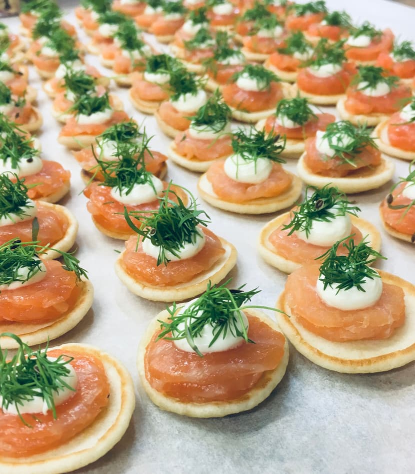 An array of smoked salmon blinis topped with crème fraîche and fresh dill, arranged in neat rows on parchment paper.