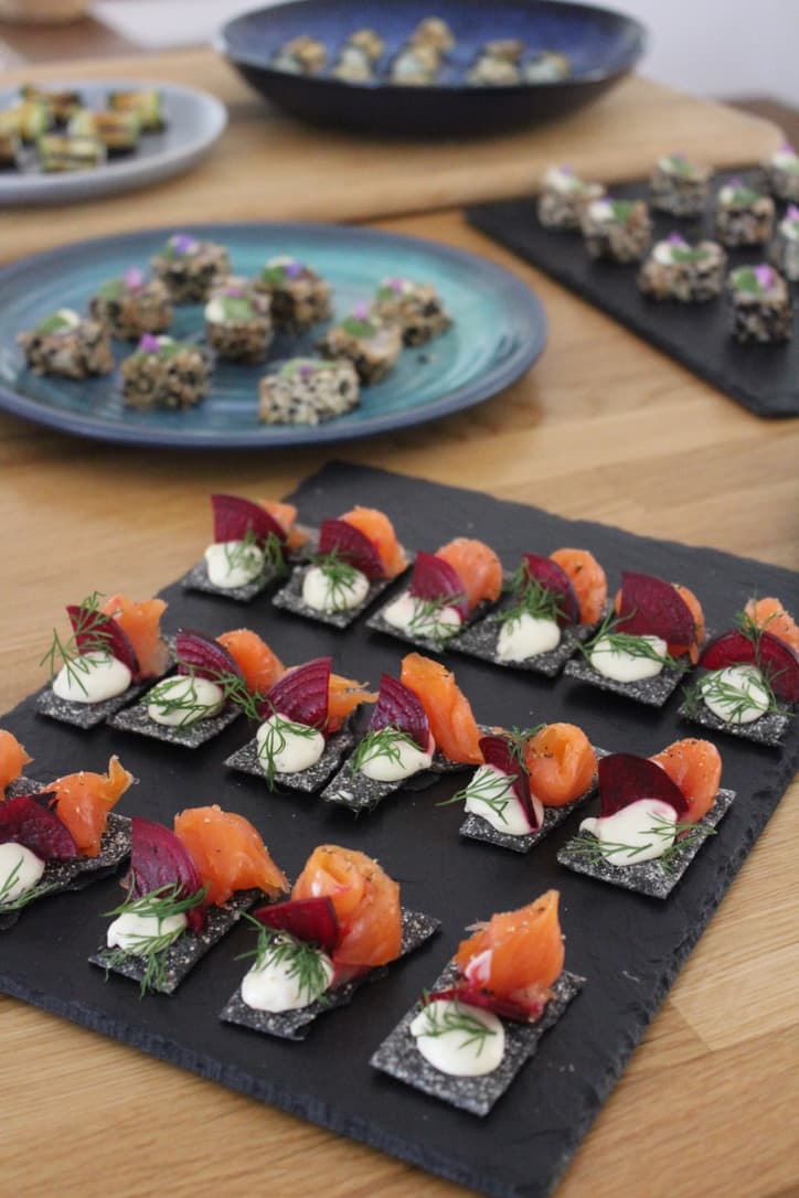 Elegant smoked salmon and beetroot canapés on black slate, garnished with fresh dill and crème fraîche, with a selection of appetizers in the background.