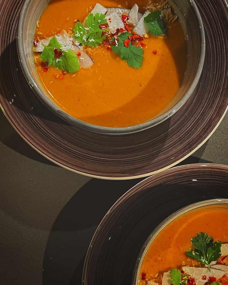 A bowl of velvety spiced carrot soup garnished with fresh herbs, coconut shavings, and red chili flakes, served in a dark ceramic bowl.